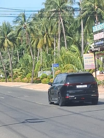 This image captures a street scene in Phu Thuy Ward, Vietnam, with a focus on a black SUV parked alongside a paved road. The vehicle has a license plate that reads "SJX-106.SFG." Behind the car, a lush tropical environment is evident, characterized by numerous palm trees with vibrant green fronds and some bare trunks. The foliage is dense, suggesting a warm and possibly humid climate.

On the left side of the image, power lines are visible, running parallel to the road and crossing over the palm trees. A sign is also present, mounted on a pole amidst the vegetation. Though partially obscured by the car and the orientation of the image, the sign appears to be advertising a restaurant, with text that includes "RESTAURANT" and some Vietnamese words. Some of the visible text on the sign appears to be: "NHÀ HÀNG REAT", "COFFEE", "BREAKFAST", and "LUNCH", indicating food and drink offerings.

The road surface is paved and appears to be in good condition, with a slight incline. The lighting suggests it is daytime, with shadows cast on the road, though the specific time of day is not definitively clear. The overall atmosphere is relaxed and tropical, consistent with a location in Vietnam. No people are clearly visible in the frame. The image has been rotated 90 degrees counter-clockwise.
