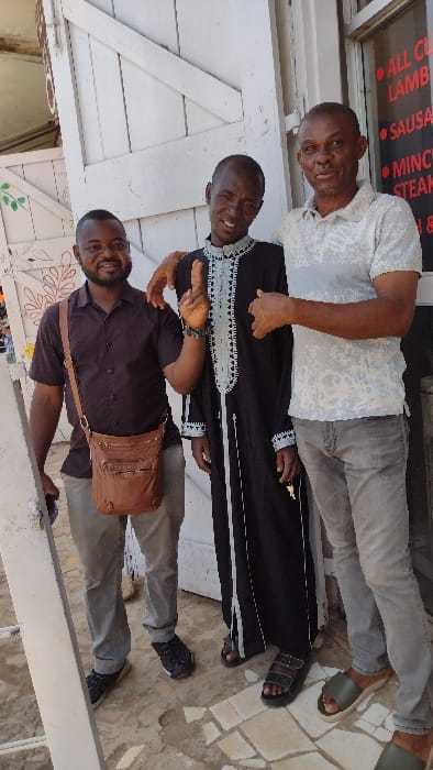 Three adult males are depicted standing outdoors in Jos, Nigeria, in what appears to be a commercial or street setting. From left to right: The first male, appearing in his 30s-40s, wears a dark brown short-sleeved button-up shirt, light grey trousers, and black sneakers. A brown leather messenger bag with multiple pockets is slung over his right shoulder. He is smiling towards the camera. The middle male, also in his 30s-40s, wears a long black traditional garment (possibly a caftan) featuring intricate white and light blue embroidery around the neckline, down the front, and at the cuffs, paired with dark sandals. He smiles broadly at the camera, his right index finger extended upwards, and his left arm resting on the shoulder of the male to his right. The third male, appearing in his 40s-50s, wears a light grey short-sleeved polo shirt with a subtle white pattern, grey jeans, and olive-green slide sandals. He offers a slight smile towards the camera, with his left arm resting on the shoulder of the middle male. The background behind the individuals includes a white wooden door or gate structure with diagonal bracing; the left portion of the door features faded green and red floral designs. To the right, a glass window displays red lettering on a white sign, listing "ALL CUTS," "LAMB," "SAUSAGE," "MINCE," and "STEAK," suggesting a butcher shop or food vendor. The ground beneath the subjects is paved with irregularly shaped light-colored tiles, possibly a mosaic of broken pieces. The scene is illuminated by bright daylight.