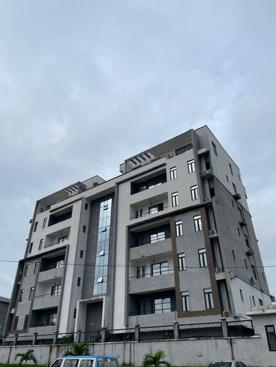 A low-angle, full shot captures a modern, multi-story building against an overcast sky in Ilado, Nigeria. The building, predominantly gray with white accents, features numerous windows and balconies. A section of the building is clad in reflective glass. The foreground shows a gray concrete wall topped with a black metal fence. Several air conditioning units are visible on the exterior of the building. A portion of a white and blue vehicle is parked in front of the fence, partially obscured by palm trees. The scene appears to be captured during daylight.