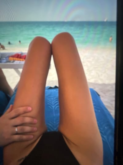 The image shows a person's legs and torso, lying on a blue towel on a beach. In the background, there is the ocean and a sandy beach with a few people. The scene appears to be a vacation setting. However, the provided location of Ravno pole, Bulgaria, is not an area known for its beaches and ocean.
