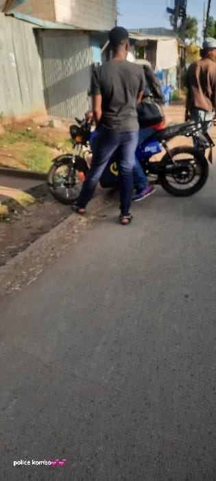 A man is pictured next to a motorcycle lying on its side on a paved street. He wears a dark long-sleeved shirt and blue jeans, facing away from the camera. Another person on a motorcycle is partially visible in the background. Buildings line the side of the road. The photo is slightly rotated clockwise. The words "police kombo" are written in pink letters in the lower right corner.