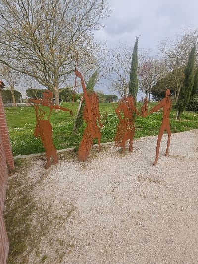 Four rusted metal cutout sculptures of human figures are positioned on a light-colored gravel path in Avignonet-Lauragais, France. The figures are arranged sequentially from left to right. The leftmost figure depicts an individual holding a horizontal object, possibly a bow or staff. The second figure has an arm raised vertically, grasping a slender, spear-like implement. The third figure exhibits patterned cutouts on its form. The rightmost figure, of a simpler silhouette, holds an object resembling a torch or club in one raised hand, extending its other arm towards the third figure.
Behind the sculptures, a landscaped area features a vibrant green lawn heavily interspersed with small white flowers. The background consists of diverse arboreal vegetation, including a large deciduous tree with nascent light-green foliage or buds, several tall, conical dark green cypress trees, and a tree displaying reddish-purple blossoms. The overhead sky is uniformly overcast and grey. A reddish-brown brick wall segment is partially visible along the extreme left edge of the foreground. Distant indistinct features are visible on the horizon.