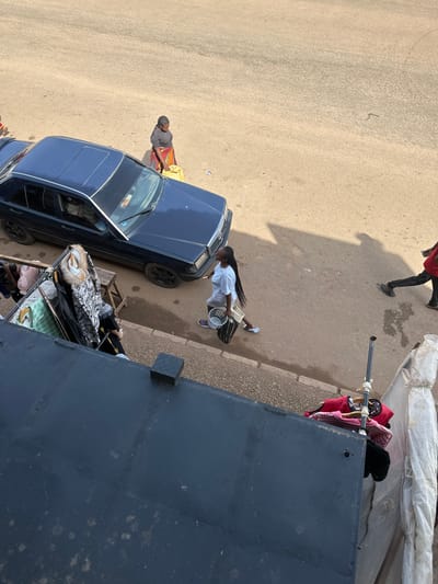 The image depicts a street scene in Bukuru, Nigeria, with several individuals and objects present. In the foreground, a dark-colored car is parked alongside a raised, dark-colored surface, possibly a building or balcony. Clothes are hung on a rack to the left of the car, and more clothing items, including a pink shirt, are displayed on a rack to the right of the car.
A woman, wearing a dark t-shirt and a colorful patterned skirt, stands near the car, holding a yellow bag. To her right, a young woman with braided hair, wearing a light-colored t-shirt and shorts, walks while carrying a white bucket and a striped bag. Further down the street, a person in a red shirt and black pants is walking away from the camera.
The street is paved with a sandy, textured surface, and shadows are cast from the car and the people onto the ground. The overall environment suggests a bustling urban or semi-urban area.