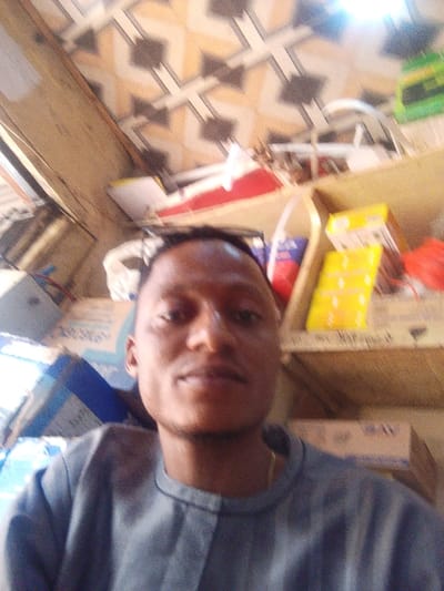 A male individual is depicted in a close-up, likely a selfie, within an indoor commercial or storage setting in the city of Ankpa, Nigeria. The subject, with dark skin and short dark hair, wears a grey, collared long-sleeved garment and a thin gold chain around his neck. His gaze is directed towards the camera. The background is moderately out of focus but reveals an active, somewhat cluttered environment. An overhead surface or wall features light-colored square tiles with a repetitive geometric pattern of dark grey and light brown elements. To the subject's right, a wooden shelving unit contains various packaged goods, including distinct yellow and red items. Below and to the left of the shelving, multiple cardboard boxes are stacked, with some exhibiting blue markings and one displaying "MAX" in visible text. The lighting appears bright, originating from an unseen source above and to the right, causing a slight glare in the upper right portion of the image. No other individuals are present.