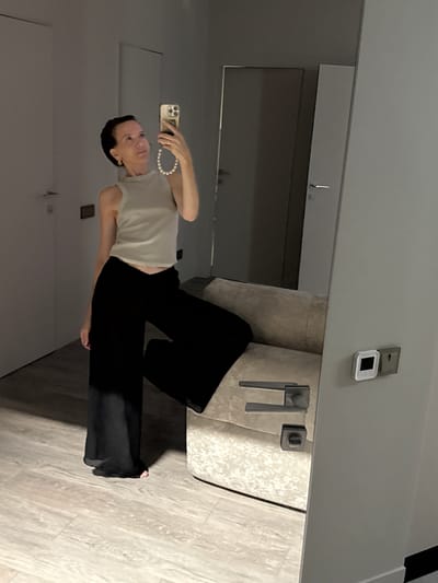 A woman is taking a mirror selfie in what appears to be a modern apartment with light-colored walls and wooden flooring. She is wearing a light-colored sleeveless top and black wide-leg pants. She has short dark hair and is wearing hoop earrings. She is holding a smartphone in her right hand, and a pearl necklace is draped over her arm. The mirror reflects a door and a piece of furniture, possibly a couch. There is a light switch and a thermostat on the wall to the right of the mirror. The image does not contain enough visual information to determine the city or country.