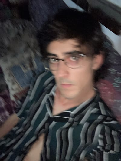 The image is a blurry, close-up shot of a person wearing glasses and a striped shirt. The person's face is the main focus, with their head tilted slightly downwards. The background is dark and indistinct, with hints of fabric or bedding. The overall impression is of a casual, indoor setting, potentially a bedroom or living area. There is no visible indication of the city of Sabadell, Spain, in the image.