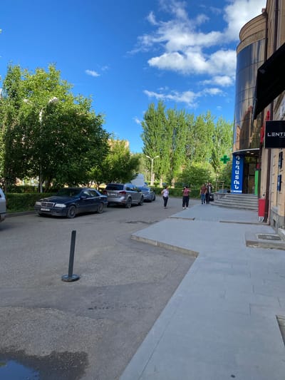 The image depicts a street scene in Gyumri, Armenia, on a sunny day with a blue sky and scattered clouds. In the foreground, a dark-colored sedan is parked on the asphalt. To the right, a modern building with a curved glass facade and an awning stands. A pharmacy with a green cross sign and text in Armenian is visible on the ground floor of the building, with several people walking towards it. Behind the cars and building, numerous tall green trees line the street, creating a verdant backdrop. The overall impression is one of a typical urban environment with a mix of residential and commercial elements.