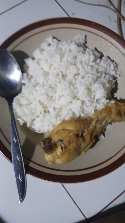 A close-up, high-angle shot shows a plate of food with a spoon, likely in a home setting in Kecamatan Pati, Indonesia. The plate, with a brown rim and a lighter beige interior, contains a mound of white rice and a piece of fried chicken. The spoon, made of stainless steel and featuring a star-like etched design on its handle, is positioned on the left side of the plate, partially submerged in the rice. The food is placed on a white tiled surface, with grout lines visible. The lighting appears to be dim, suggesting an indoor environment possibly at night.
