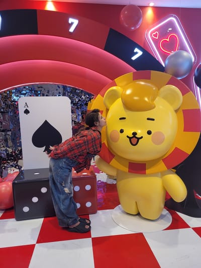 A person wearing a red and black plaid shirt and distressed denim jeans is leaning in to kiss a large, yellow cartoon lion statue. The lion has a mane of red and yellow stripes. The statue is part of a colorful, casino-themed display. Behind the person and to the left, there is a giant Ace of Spades playing card. Two oversized dice, one black and one red, are positioned in front of the card. The background features red arches, illuminated numbers "7", a neon heart playing card, and suspended red and silver spheres. The floor is a red and white checkerboard pattern. The scene appears to be located in a vibrant, indoor entertainment venue, likely a themed attraction or exhibition, possibly in Busan, South Korea, given the context of the prompt.