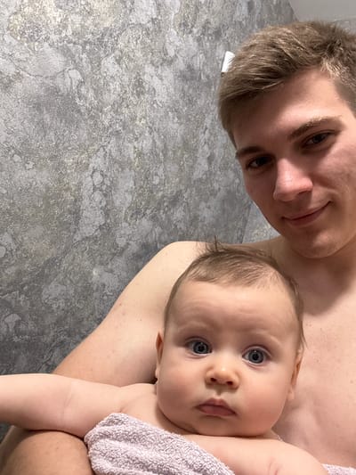 A young Caucasian man, appearing to be in his early twenties, is holding a baby. The man has short, light brown hair and is shirtless. He is looking towards the camera with a slight smile. The baby, with blue eyes and light brown hair, is positioned in front of the man, also looking towards the camera. The baby is wrapped in a light purple towel. The background is a textured wall with a grey and metallic finish. The image appears to be a selfie taken indoors. There is no indication of the specific location within Krasnoyarsk, Russia.