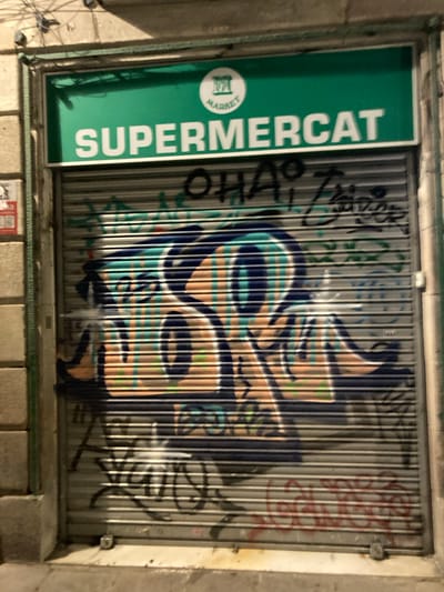 The image shows a closed metal security shutter on a storefront in Barcelona, Spain. The shutter is adorned with graffiti. Above the shutter, a green sign reads "SUPERMERCAT" in white capital letters, with a circular logo containing the word "MARKET". The building's facade is made of stone or concrete. The lighting suggests it is nighttime or an interior setting.