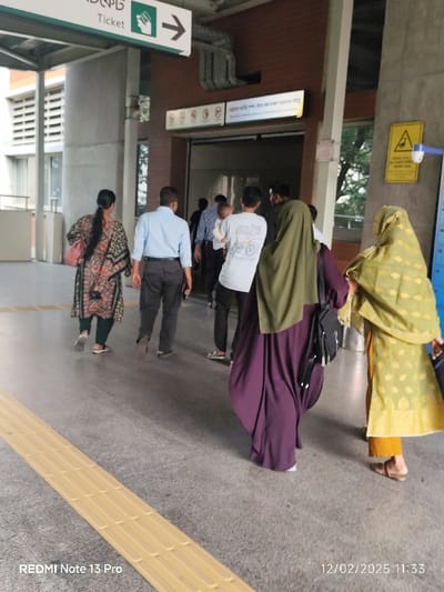 Several people walk through what appears to be a transportation hub. To the left, a sign points towards a ticket booth. Individuals are dressed in various styles, including casual clothing, office attire, and traditional Muslim garments. The scene is well-lit, with a focus on the people moving towards an entrance or exit. A textured yellow strip runs along the floor, likely for accessibility. A warning sign is visible near a doorway. The photograph includes a timestamp at the bottom.