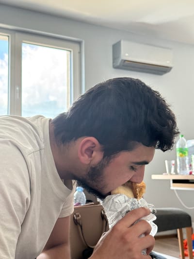 This media file captures a side-profile portrait of a young man, presumably in Sofia, Bulgaria, intensely focused on eating a large sandwich or burger. He has dark, curly hair and a short beard, and is wearing a light-colored, possibly beige, t-shirt. His eyes are closed as he takes a bite, indicating a moment of concentration or enjoyment. The food is partially wrapped in white paper, which appears to have some indistinguishable text or pattern printed on it.

The setting is an indoor space, brightly illuminated by natural light from a large window on the left side of the frame, revealing a bright sky with some clouds, suggesting it is daytime. On the upper left wall, a white, modern air conditioner unit is visible. The background also shows glimpses of blurred furniture and plastic bottles, indicating a casual indoor environment, possibly a home or office. The man's right arm and hand are visible as he holds the food. There is no legible text visible in the image.