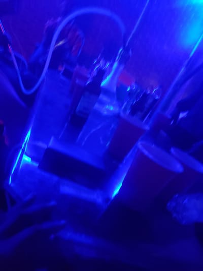 The scene depicts an indoor environment within the city of Barden Yana, Nigeria, characterized by heavy blue lighting and significant motion blur. The setting appears to be a nightclub or bar, operating in low ambient light. A prominent white, glowing, serpentine tube-like structure extends across the upper and central portions of the frame. Below this, multiple dark, reflective surfaces, possibly tables or bar tops, are visible, some displaying red or orange undersides or elements. Indistinct bottles, glasses, and red cups are discernible on these surfaces. Blurred human figures are faintly present in the background and mid-ground, some exhibiting reddish tones on their heads or upper bodies. Bright streaks of blue light emanate from various light sources, illuminating the dynamic and obscured environment.