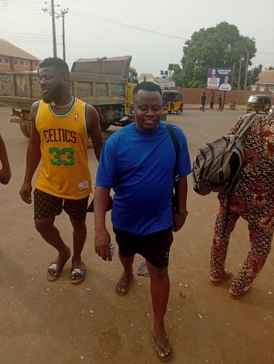 Three men are captured walking outdoors on what appears to be a street or unpaved pathway in Onitsha, Nigeria, under an overcast sky.
In the foreground, a man on the left, with a beard, wears a yellow basketball jersey with "CELTICS" and the number "33" printed on it, black and white patterned shorts, and grey Nike slides. He looks towards his left. Next to him, a man in a blue t-shirt, black shorts, and flip-flops walks towards the viewer's right, carrying a black shoulder bag. His expression is relaxed, with a slight smile. Partially visible on the right is another person, seen from the waist down, wearing brown and red patterned trousers or a skirt and carrying a grey backpack. This person also wears flip-flops.
The background reveals a bustling urban scene. A large, worn dump truck and another truck are parked or driving in the mid-ground. Further down the road, two yellow tricycle taxis, known as Keke Napep, are visible, along with several other people walking. Power lines and utility poles crisscross the top of the image. On the right side, a billboard displays text "LIFE OF FAITH" and "LUMEN CHRISTI" alongside a portrait of a man. The ground is a mix of dirt and possibly worn asphalt.
The image suggests a daytime setting, possibly morning or afternoon, given the natural light and the activity. The overall mood appears casual and everyday.