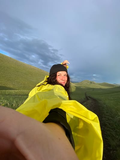 A woman wearing a black beanie and a bright yellow hooded rain jacket is looking back at the camera while holding up a part of the jacket. She is in a grassy, hilly landscape with a dirt path leading into the distance. The sky is cloudy with patches of blue and sunlight. The scene appears to be outdoors in a natural environment, possibly near the city of Verkhneozerskoye, Russia, given the rolling hills and vegetation. There are a couple of other figures visible on the path in the distance.