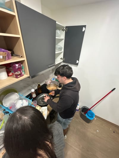 The scene depicts an interior kitchen located in Bragança, Portugal. Two individuals are present. A male, with short dark hair, wearing a dark hoodie and light-colored shorts, stands at the center-right, facing left. He is actively engaged in cooking, holding a metallic utensil over a frying pan that contains a light-colored residue, positioned on a stovetop. Adjacent to the frying pan is a smaller pot holding a light brown liquid. A brown bottle and a red bottle with a white cap are situated on the counter behind the cooking implements. Above the cooking area, dark grey overhead cabinets are mounted; one cabinet door is open, revealing white interior shelves containing glassware. To the left, a female individual is partially visible in the foreground, seen from behind, with long dark hair and wearing a grey textured garment. On the counter to her left, a blue plastic dish drying rack holds several white dishes. Open wooden shelving units are located above the drying rack, containing various packaged goods. The kitchen walls are uniformly white, and the floor is composed of light-toned wooden planks. In the lower right corner, a grey mop bucket with a blue wringer and a red-handled mop are positioned against the wall. A white plastic bottle is also visible near the bucket.