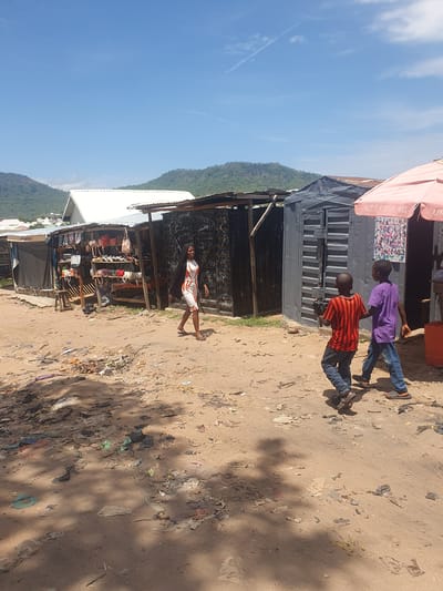The image captures an outdoor scene, likely a market or street, in what appears to be a semi-urban or rural area, with a prominent mountainous landscape in the background under a clear blue sky. The ground is a dirt path littered with various debris and casting distinct shadows, indicating a bright, sunny day.
In the foreground, a young woman or older girl stands barefoot on the left side of the dirt path, wearing a white dress with orange and red geometric patterns. Behind her, market stalls are visible, constructed from materials like corrugated metal and wood, with various goods, including an array of handbags, displayed on shelves.
On the right side of the dirt path, two young males, likely children or teenagers, are walking. The one in front wears a red and black striped t-shirt and blue jeans, while the one behind him wears a purple t-shirt and blue jeans. They are walking away from the viewer, moving towards the background.
Above the market stalls, a red awning is partially visible, with text that reads "GET FREE 4G" and another partial word, possibly "UP." A poster depicting multiple faces is affixed to the wall of one of the stalls beneath this awning. The scene depicts daily life and commerce in an outdoor setting.