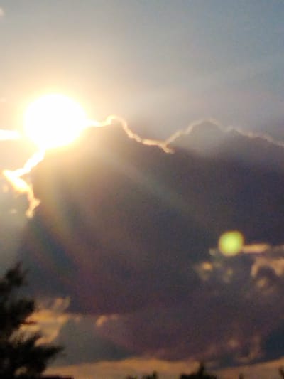 A sunset is depicted in Ustyvytsia, Ukraine. The sun, a bright, luminous orb, is positioned in the upper left quadrant of the image, partially obscured by dark, dramatic clouds. Rays of light emanate from the sun, creating a dramatic backlight effect. The clouds are varied in shape and density, with some appearing dark and heavy, while others are illuminated by the sun. In the lower left of the image, the silhouette of trees or foliage is visible. The sky transitions from a pale blue at the top to darker hues closer to the horizon. A blurry, yellow-green circular object is present in the middle right of the image, possibly a lens flare or another celestial body. The overall impression is of a powerful and somewhat turbulent natural phenomenon.