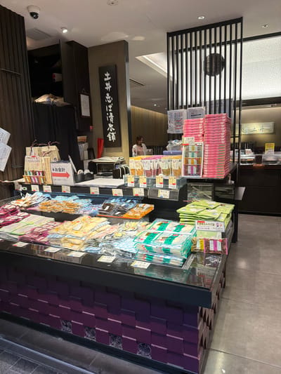 The image shows the interior of a shop in Kyoto, Japan, displaying a wide variety of packaged food items, likely souvenirs or local specialties. The products are arranged on refrigerated display shelves and on tiered stands. Many of the packages are in plastic wrapping and feature colorful designs and Japanese text. Some of the items appear to be dried goods, snacks, or confectionery.

In the background, there is a vertical sign with Japanese calligraphy. A woman is visible in the background, appearing to be a staff member, standing behind a counter where there is a computer monitor and other retail equipment. The shop has modern fixtures, including a black metal partition with vertical slats and a white patterned screen behind it. The overall lighting suggests an indoor retail environment. The flooring appears to be a grey concrete or tile. The display units have purple decorative paneling at their base with visible cooling fans. A red and white sign with an arrow pointing to the right, indicating "Payment" or "Checkout" in Japanese, is prominently displayed on a shelf.