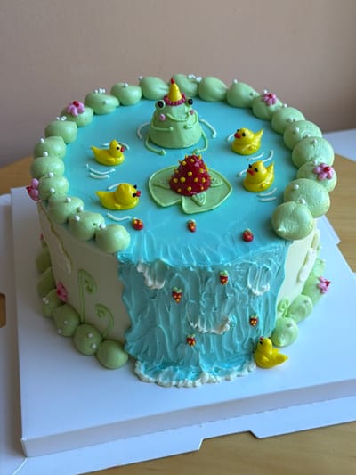 The image displays a creatively decorated cake with a pond and frog theme. The cake has a light-colored frosting on its sides, adorned with green, bulbous piped frosting elements that resemble lily pads or rounded leaves, interspersed with small pink floral accents. The top of the cake is covered in a light blue frosting, simulating water.
Dominating the "water" surface are several yellow, piped ducklings. In the center, a green frog wearing a yellow and pink party hat sits on a green lily pad. Next to the frog, another lily pad holds a red, strawberry-shaped decoration with yellow dots. Smaller red and yellow strawberry decorations are scattered across the blue frosting.
A significant feature of the cake's side is a textured blue frosting waterfall cascading down, with white frosting forming the frothy base. More small strawberry decorations are visible on and around the waterfall. The cake is presented on a white cake board.
The background of the image is a plain, light beige wall and a wooden surface, suggesting the cake is indoors, possibly on a table. There is no indication of a specific location in Tel-Aviv, Israel, or any people present.