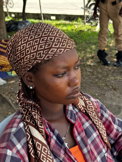 The image depicts a dark-skinned woman with her head and upper torso visible. She is wearing a brown and white patterned headscarf and a red and blue plaid shirt over an orange top. She has jewelry in her ear. The background shows some blurred vegetation, pavement, and the lower legs of at least one person wearing brown trousers and black shoes. The location is Itirin, Nigeria.