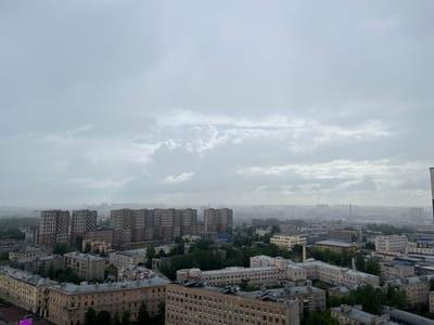 This is an aerial, eye-level, medium shot of a dense urban environment on a cloudy day, likely in Imeni Morozova, Russia, given the architectural style of the buildings and the overall atmosphere. The scene is dominated by a multitude of apartment buildings of varying heights and architectural periods. In the foreground, older, lower-rise buildings with light-colored facades and grey roofs are visible. Behind these, several taller, modern apartment blocks with a patterned facade stand out. The buildings are interspersed with a significant amount of greenery, primarily trees, indicating a well-established residential area with parks or tree-lined streets. The background shows a sprawling cityscape fading into a hazy distance, suggesting a large urban area. The sky is heavily overcast with thick grey clouds, creating a muted and somber light across the entire scene. There are no visible people or vehicles, and the environment appears quiet and still. The overall impression is one of a vast residential district under a gloomy sky.