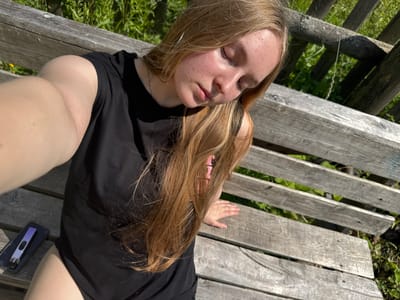A young person with blonde hair is lying on a wooden bench, taking a selfie in the sunlight. They are wearing a black t-shirt. To their right, on the bench, is a dark case containing a light purple electronic device. The bench is outdoors, with green grass and foliage visible below and around it, suggesting a natural or park-like setting. The lighting indicates it is daytime, with the sun casting shadows and highlighting the subjects. The person's eyes are closed, and their expression appears relaxed, possibly enjoying a moment of repose. The presence of the electronic device could imply they are pausing from their activities or simply relaxing with their belongings. The scene is set in Ryabinino, Russia.