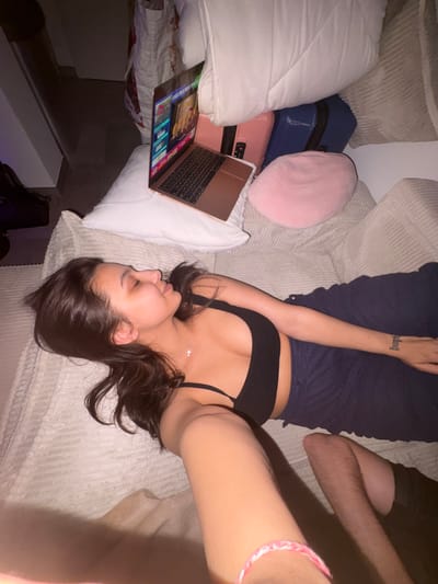 In Mönchengladbach, Germany, a young woman is seen lying on a couch, resting her head on a pillow. She is wearing a black top and dark pants. A laptop is open on a stack of luggage next to her, displaying content. A pink cushion is also visible on the luggage. The room appears to be dimly lit.