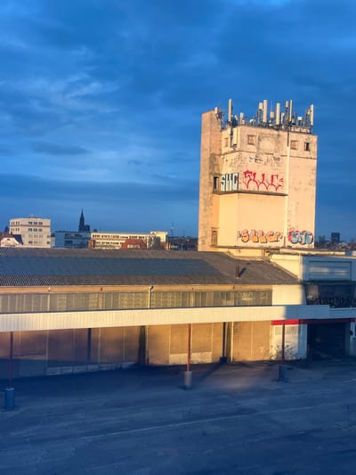 An urban landscape shows several buildings under a bright sky. A tall, weathered building with antenna arrays and graffiti on its facade dominates the left. Adjacent is a modern building with long windows and a white stripe detail. Other buildings are visible in the background, including one with a distinctive spire. The foreground features a flat, blue area with what appear to be ventilation shafts.
