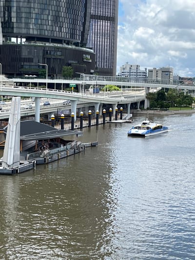 This image captures a vibrant urban river scene, likely during the daytime under a partly cloudy sky. The primary setting is a wide river, with an active city waterfront on the left bank.

In the foreground and to the left, tall, modern buildings with extensive glass facades define the urban skyline. In front of these structures, an elevated concrete walkway or bridge, supported by sturdy grey pillars, curves along the river's edge. Beneath this elevated pathway, a long, dark-colored floating pontoon or ferry terminal extends along the bank. This pontoon features several black bollards with bright yellow tops and a covered area, suggesting a waiting or boarding point. A white sign on the pontoon is partially visible and clearly displays the text "NORTH QUAY," indicating a specific location along the Brisbane River.

On the river itself, a sleek, blue and white catamaran-style boat, characteristic of a public transport ferry or water taxi, is visible. It appears to be moving, creating gentle ripples in the otherwise calm, light brown water. The boat is positioned in the mid-ground, heading away from the pontoon towards the right side of the frame. The scene is one of everyday urban activity, showcasing the city's infrastructure and river transport system.