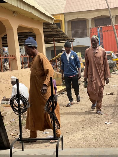 An outdoor scene in Zaria, Nigeria, depicts a busy, unpaved street or market area under daylight conditions. In the left foreground, an older man, wearing a brown long-sleeved traditional garment and a dark blue cap, stands in profile, facing right. He holds a thin object between the fingers of his right hand. Directly in front of him is a black metal display rack holding multiple coils of black wire. To his left and slightly behind, a man in a white garment is seated on a low structure.
In the midground, moving from left to right, a younger man walks, wearing a blue zip-up hoodie with a white v-neck shirt underneath, black pants, and black slides. He also wears a black beanie with a knot at the top. To his right, an older man walks in the same direction, dressed in a light brown long-sleeved traditional garment and a patterned green and yellow cap. He wears blue flip-flops and carries two green, possibly plastic, containers in his left hand.
The ground is predominantly dirt or dusty earth, with scattered debris including white litter. Buildings line the background, featuring a mix of painted structures, some with corrugated metal roofs. A yellow tricycle (Keke Napep) is visible in the far right background, adjacent to a large red shipping container. The architecture suggests a commercial or residential district.