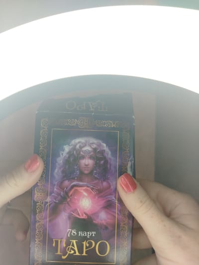 A pair of hands, likely a woman's, with short, light red/pink nail polish, holds a dark blue or purple rectangular box, presumably for a deck of tarot cards. The box is adorned with gold decorative borders and features a central illustration of a mystical woman. She has voluminous, light-colored, curly hair, wears a decorative headband, and has bracelets on her wrists. Her hands cradle a glowing, ornate orb, and her attire is depicted in flowing shades of purple and pink.
Visible text on the box front reads "78 карт" (meaning "78 cards" in Russian) and "ТАРО" (meaning "Tarot" in Russian). The top flap of the box also shows the mirrored and upside-down text "ТАРО". The entire scene is brightly lit from above by a prominent ring light, which causes significant overexposure and glare in the upper part of the image, obscuring details beyond the box and hands.
The setting is indoors, with the strong artificial light suggesting a staged photograph or video recording. No specific time of day, weather, or emotions are discernible from the image itself, but the Cyrillic text is consistent with the provided location context of Izhevsk, Russia.