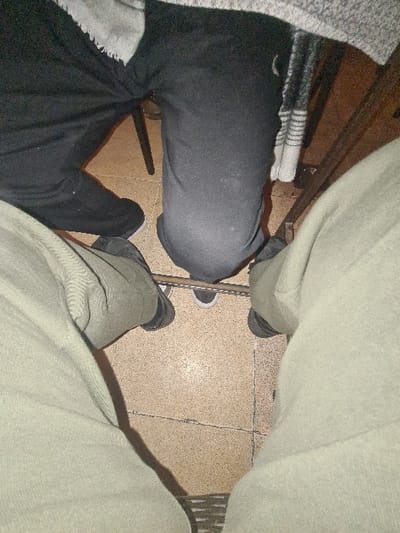 A low-angle shot captures two seated figures with distinct clothing styles. On the left, black pants and a gray sweater are visible, while the figure on the right wears khaki pants. The flooring is composed of square tiles. Shadows suggest a light source above. The unusual perspective focuses primarily on the lower body and highlights contrasting textures and colors.