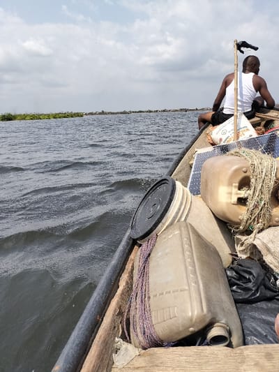 The image depicts a wooden boat navigating a waterway, identified as likely the Lagos Lagoon or an interconnected channel in Lagos, Nigeria, under a partially cloudy sky. An adult male, wearing a white undershirt and dark shorts, is seated at the stern, facing forward. The boat's interior is loaded with various items: two light brown plastic jerrycans, one secured with purple rope, a black plastic bag, a dark flexible solar panel, a partially visible white sack, and a stack of plastic buckets topped with a black lid. A slender wooden pole with an attached dark object rises vertically behind the seated man. The dark water surface shows ripples, indicating movement. To the mid-left, a patch of green aquatic vegetation is visible. The distant horizon reveals a low-lying settlement composed of multiple structures along the shoreline.