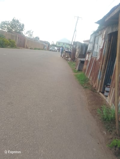 The image shows a street scene in Jos, Nigeria. The road is asphalt and occupies most of the foreground. The right side of the street is lined with structures made of corrugated metal, possibly shops or dwellings. Vegetation grows between the road and these structures. A power pole is visible further down the street. In the distance, there is a car and a few people standing near a building. On the left side of the road is a concrete wall and building. The sky is overcast. "Q Express" is printed in the bottom left corner.
