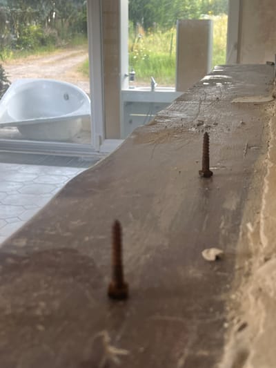 The image shows two screws standing upright on a concrete surface. The surface appears to be a ledge or countertop. In the background, through a large window, a bathtub is visible, as well as a grassy outdoor area with trees and a dirt road. The scene suggests an unfinished construction or renovation project. The specific location in Dilbeek, Belgium, is not provided by the image itself.