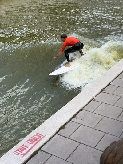 A surfer, wearing a bright orange top and black leggings, navigates a standing wave in a river in Rotterdam, Netherlands. The surfer is on a white surfboard, with the tail of the board angled upwards as they ride the wave. The water is choppy and turbulent, creating a white spray around the surfer.
The scene is viewed from above, looking down at the river and the adjacent bank. The bank is paved with light gray rectangular stones, bordered by a concrete edge. The concrete edge has red spray-painted text that reads "STAFF ONLY!". Green grass and small weeds grow in the cracks between the paving stones and along the edge of the concrete.
The lighting suggests it is daytime, likely sunny, with clear reflections visible on the water's surface. The overall atmosphere appears energetic, focused on the activity of surfing. The combination of the urban setting with the river and the surfing activity creates a unique urban watersport scene.