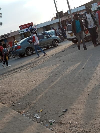 The image depicts a street scene in Ildamat ward, Kenya, during daylight hours, characterized by long shadows suggesting late afternoon or early morning. The foreground features an unpaved, light-brown dirt road with tire tracks and scattered refuse including plastic wrappers, a small plastic bottle, and other debris. A concrete curb runs along the lower left edge.

Several individuals are present on the road. From left to right:
1.  A person, partially visible, wearing dark clothing and carrying a backpack, walks from right to left.
2.  A male individual walks from left to right, wearing a red and white jacket, blue jeans, and striped sandals. He carries a bundle of white, possibly fabric, items in his left hand and balances a larger, similar bundle on his head.
3.  A male individual walks from left to right, wearing a brown jacket, a light blue t-shirt, dark trousers, and dark shoes.
4.  Another male individual walks from left to right, wearing a patterned jacket, a white shirt, dark trousers, and dark shoes. He holds a black mobile phone to his right ear.
5.  A fifth person is partially visible behind and to the right of the individual on the phone, wearing a red top.

Vehicles are present on the road. A blue-green sedan with dark wheels is positioned behind the individual carrying items on his head. Further to the right, a white SUV or van is visible, displaying a Kenyan license plate "KCA 7 197D" on its rear, partially obscured. A motorcycle with a rider is visible behind the white vehicle.

In the background, a line of single-story commercial buildings runs parallel to the street. Visible signage includes "FASHIONS" on the left, "M-PESA EMAN" and "EKALA CHEMSIST" beneath a red Coca-Cola sign in the center-left, and a partially visible sign possibly reading "BUTCHERY" further right. Utility poles with multiple overhead wires are prominent on the right side of the frame.