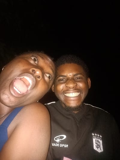 Two people are visible in a low-light selfie. A woman on the left has her mouth wide open, eyes looking up, and is wearing a blue top. A man on the right is smiling broadly, showing his teeth, and has a neatly trimmed beard. He is wearing a black shirt with a logo on the left side and a square emblem on the right. The background is dark and mostly obscured.