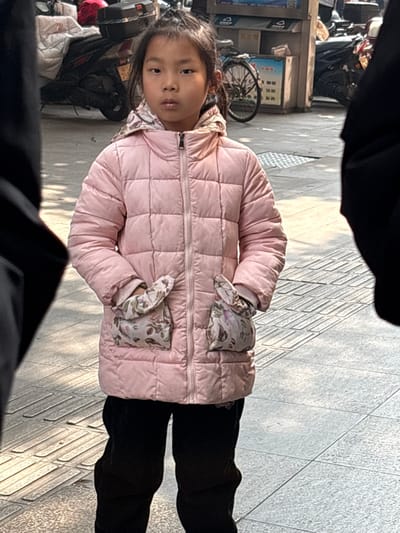 A young girl wearing a pink quilted jacket with floral patterned gloves stands on a sidewalk. She has dark hair and a serious expression. Parked motorcycles and a bicycle are visible behind her, suggesting an urban setting. The sidewalk has a textured surface, and the overall lighting indicates daytime. Dark silhouettes of people are at the top and bottom of the frame, likely the photographer.