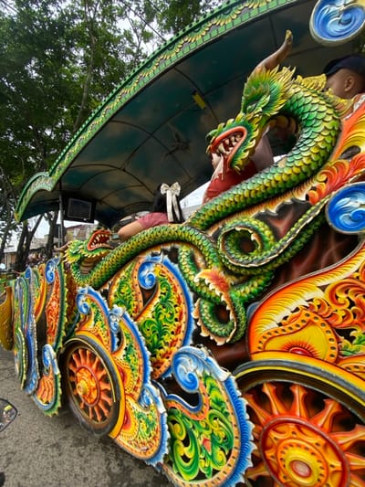 A vibrant, ornate structure features a green and yellow dragon with an open mouth, its scales intricately detailed. Adjacent to the dragon are stylized wheels and floral motifs, rendered in bright oranges, yellows, and blues. The background includes a glimpse of a dark green canopy, a person wearing a hat, and tree branches against a cloudy sky. The overall effect is of a colorful, elaborately decorated vehicle or architectural element.