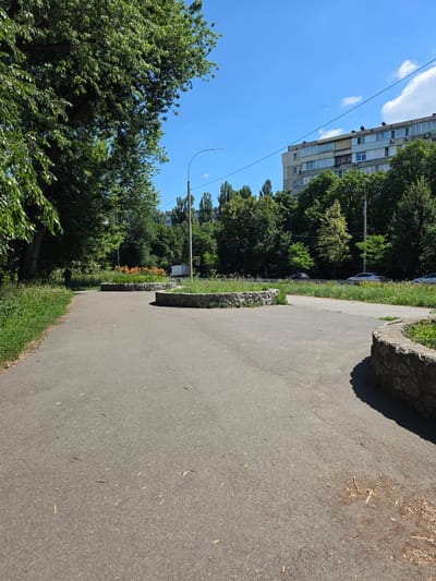 The image depicts a street scene in Kyiv, Ukraine, on a sunny day. A paved pathway runs diagonally across the frame, bordered by lush green grass and trees. In the background, a multi-story building stands tall, with windows and balconies visible. A line of trees separates the building from the pathway, and beyond that, a clear blue sky with a few wispy clouds suggests fair weather.
The pathway itself is made of asphalt and shows signs of wear, with some small twigs and debris scattered on its surface. A large, rough-hewn stone sits on the edge of the pathway, casting a shadow. Further along the pathway, a flowerbed with vibrant orange flowers adds a splash of color to the scene. A lamppost is also visible, suggesting the presence of street lighting.
The overall impression is one of an urban environment that is well-maintained, with green spaces integrated into the cityscape. The bright sunlight and clear sky indicate it is daytime, likely late morning or early afternoon. There are no people visible in the image, giving it a tranquil and serene feel. The composition, with the pathway leading the eye into the scene, invites the viewer to imagine a walk through this pleasant urban setting.