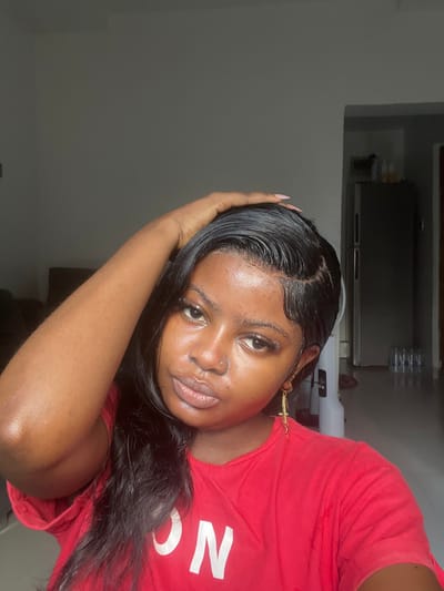 The image features a single female subject of young adult age, positioned indoors, gazing directly into the camera. She has a dark complexion, long dark hair styled with visible "baby hairs" along the hairline, and wears a red short-sleeved t-shirt with partial white lettering on the lower left. Her right hand is raised, fingers touching the top of her head near the left temple, revealing light pink nail polish. A small piercing is present on her right nostril, and a gold-colored cross dangle earring hangs from her right ear. The background shows a residential interior with light-colored walls and a lighter-toned floor. To the subject's right, a silver-colored two-door refrigerator/freezer unit is visible, with multiple plastic bottles containing clear liquid stacked on the floor adjacent to it. An indistinct dark object, possibly furniture, is partially visible on the far left. The lighting is moderate and natural, illuminating the subject and the immediate surroundings. No discernible features within the image explicitly identify the location as Badore, Nigeria.