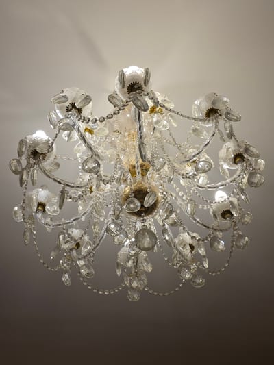 A crystal chandelier hangs from a white ceiling, casting light and shadows. The chandelier features multiple tiers of clear glass or crystal droplets and faceted beads, connected by delicate chains. Some of the glass elements are shaped like flowers, with brass-colored accents at their centers. The central part of the chandelier has a golden, possibly brass, fixture. The overall impression is one of ornate, traditional lighting. The image provides no information about the city of Veliky Novgorod, Russia, or any people, actions, or interactions. The environment is limited to the ceiling and the chandelier itself.