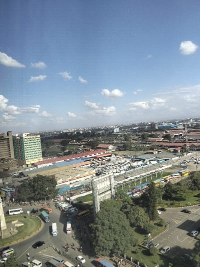 This is an aerial view depicting a bustling urban landscape in Nairobi, Kenya, under a clear blue sky with scattered white cumulus clouds.

In the mid-ground and foreground, a complex infrastructure of roads, buildings, and transport hubs is visible. To the far left, a tall, multi-story building with distinctive green-paned windows, identified as Afya Centre, stands prominently. Adjacent to it on the left is another tall, darker-colored building. Below these structures, numerous single to multi-story commercial buildings with roofs of varying colors (red, blue, yellow, grey) line a main thoroughfare.

A large, circular roundabout with a central, light-colored monument or obelisk structure is visible in the lower left quadrant. Multiple vehicles, including white vans, blue and white buses, motorcycles, and dark cars, are navigating or parked along the roundabout and adjacent roads. Pedestrians are sparsely visible on sidewalks and crossing areas.

The central and right-hand mid-ground is dominated by a large bus station or terminus, characterized by numerous parked and queued public transport buses of various colors (yellow, blue, red, white). This area features parallel elevated structures, possibly railway lines or pedestrian walkways, and extensive concrete and asphalt surfaces. A large, multi-panel billboard frame with a white support structure is centrally located, partially obscuring the bus terminal from the foreground.

Extensive patches of mature green trees and manicured lawns are interspersed between the roads and the bus terminal, particularly in the lower right and mid-ground. Roads in the lower right show additional parked cars.

The background consists of a dense urban sprawl extending to the horizon, with numerous buildings of varying heights and types, fading into a hazy distant skyline. The overall scene suggests a vibrant commercial and transportation hub within Nairobi's Central Business District, likely observed from an elevated vantage point overlooking areas such as the Country Bus Station and surrounding commercial streets.