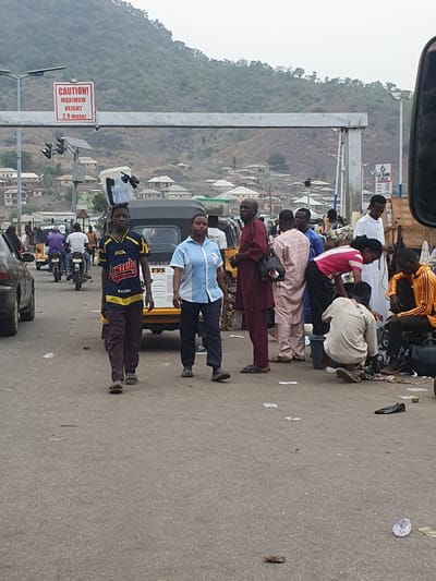 The image depicts a bustling street scene in Lokoja, Nigeria, characterized by human activity and vehicles under an overcast or hazy sky. Numerous people, mostly adults, are congregated along the side of a paved road, suggesting a bus stop, market, or common gathering area.
In the foreground and midground, individuals are seen standing, walking, and some are seated on low stools or directly on the ground. A prominent figure in a blue and yellow t-shirt with the word "Money!" printed on it stands in the midground, while another man in a light blue shirt is positioned near a yellow and black auto-rickshaw (tricycle). Motorcycles and other vehicles are visible further down the road, indicating active traffic flow.
The background features a hilly landscape with sparse vegetation, upon which numerous residential buildings are scattered. A large metal structure spans across the road, likely a support for overhead signage or a pedestrian bridge. Attached to this structure are traffic lights and a visible white sign with red text that clearly states: "CAUTION! MAXIMUM HEIGHT 2.9 meter". Small solar panels are also mounted on poles near the traffic lights, possibly powering them. The road surface shows some scattered debris. The scene conveys the busy, everyday life of an urban environment in the region.