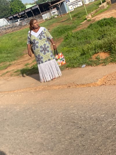 A middle-aged Black woman with braided hair walks along a dusty path next to a road. She is wearing a long, patterned dress with a white, v-shaped neckline and a fringed hem. In her right hand, she carries a multi-colored tote bag. The background shows a grassy area with some scattered white flowers, a sign for "KWANGAH DRIVING SCHOOL," and several structures, possibly part of a market or workshop, with corrugated metal roofing and wooden fencing. The overall scene appears to be in an outdoor, possibly semi-rural, setting within the city of Jos, Nigeria, suggested by the presence of local businesses and vernacular architecture.
