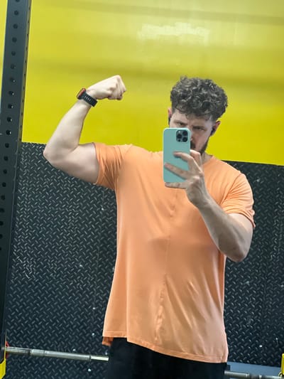 A man with curly dark hair and a beard is taking a mirror selfie in a gym. He is wearing an orange t-shirt and black pants. He is flexing his right bicep and holding a light blue smartphone in his right hand, angled to capture his reflection. He also appears to be wearing black earbuds. The background of the gym features a bright yellow wall and a black diamond plate textured wall. A metal weight rack and a barbell are partially visible on the left and bottom edges of the frame respectively. The overall scene is consistent with a fitness facility.