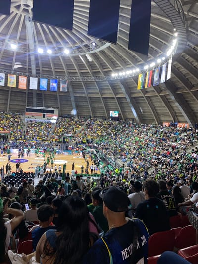 This image captures a packed basketball game inside a large, dome-shaped arena. The arena features a distinctive wooden, ribbed ceiling structure with numerous spotlights illuminating the court. Banners are hung from the upper levels of the arena.
The court itself is visible, with players in green uniforms engaged in a game. Spectators fill the tiered seating surrounding the court, many wearing green, suggesting they are supporting the home team. The crowd is dense, with people of various ages and ethnicities.
In the foreground, spectators are seated, some in focus and others blurred. One individual in the immediate foreground wears a blue and yellow jersey with "NEYMAR JR." printed on the back. Other individuals in the foreground are looking towards the court or at their phones.
The atmosphere appears energetic and lively, indicative of a sporting event. The overall scene depicts a community event centered around a basketball game in a significant indoor venue within the city of La Asuncion, Venezuela.